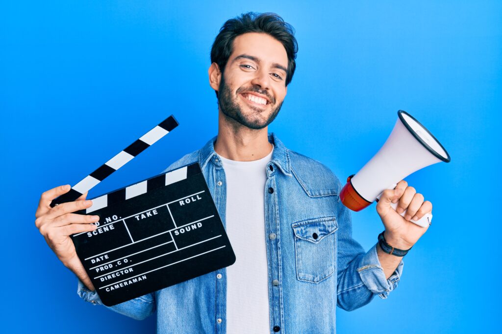 young hispanic man holding video film clapboard and megaphone smiling with a happy and cool smile on face. showing teeth.